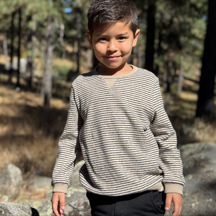 A young boy standing outdoors in a wooded area, wearing a beige and gray horizontally striped long-sleeve shirt and dark pants.