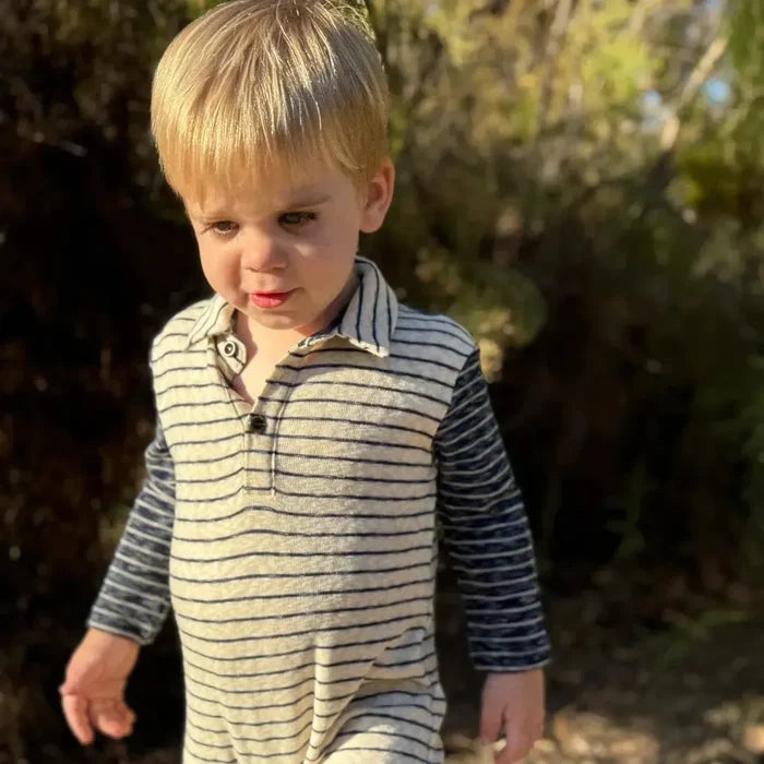 A young blonde boy wearing a striped long-sleeve footless romper jumpsuit walks outdoors on a dirt path with green bushes in the background.