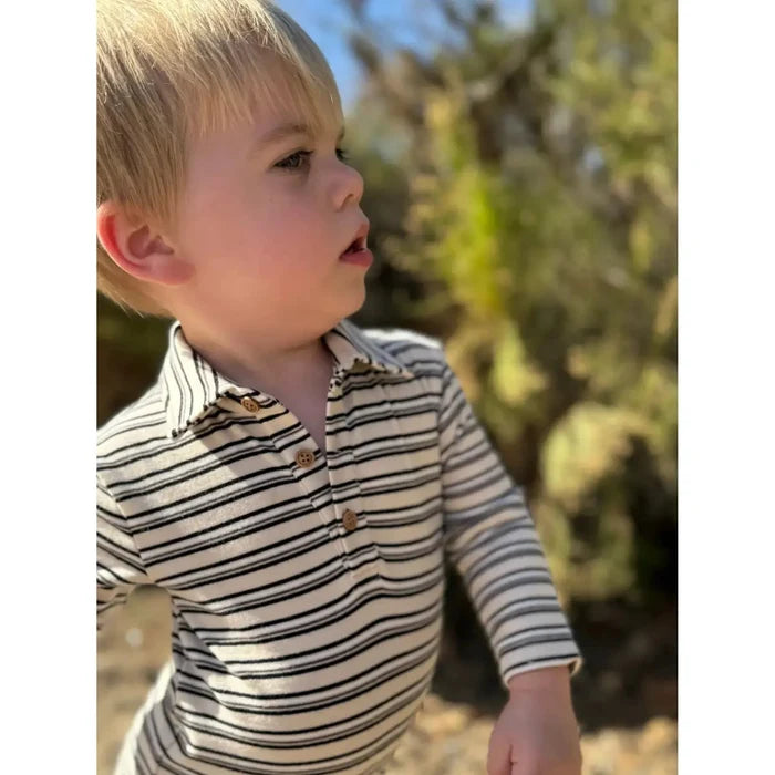A young blonde boy wearing a cream long-sleeve polo shirt with thin black horizontal stripes and brown buttons, photographed outdoors with a blurred natural background.