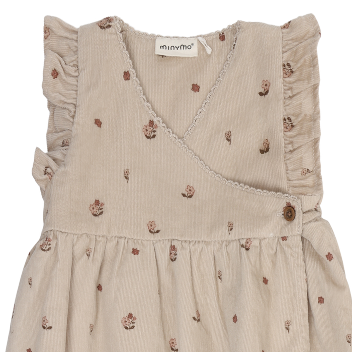 A sleeveless beige baby dress with a small floral print, featuring ruffled shoulder details and a wrap-style V-neckline, shown against a plain white background.