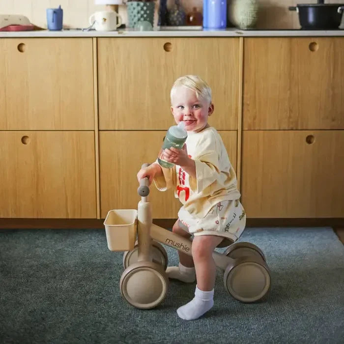 Child sitting on a wooden tricycle in a kitchen holding a green bottle.