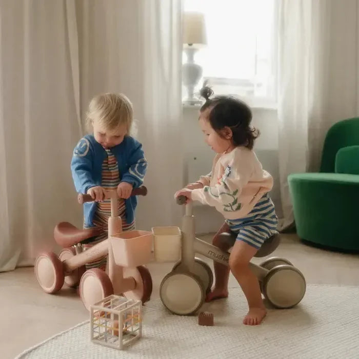 Two children playing with wooden balance bikes in a room with curtains and a green chair.