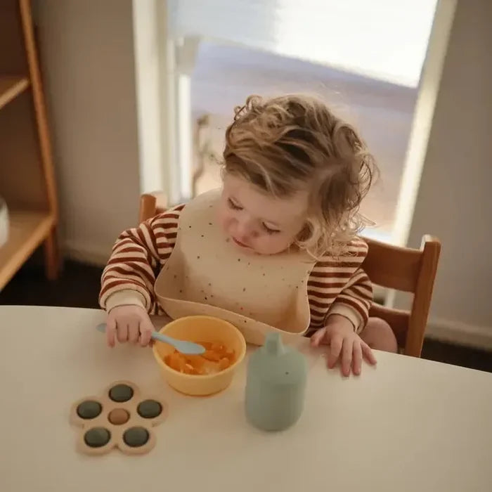 Child sitting at a table with a bowl of food, a spoon, and a bottle.