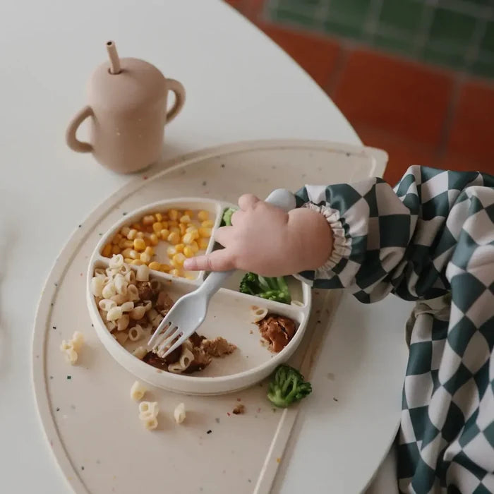 Child's hand reaching for food on a divided plate with a cup in the background.