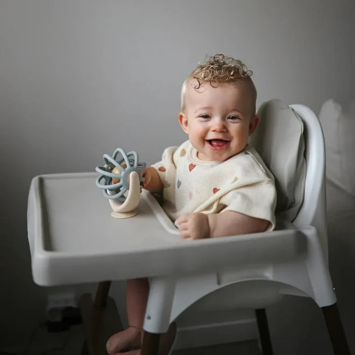 Baby sitting in a high chair with a spinng ball toy, smiling.