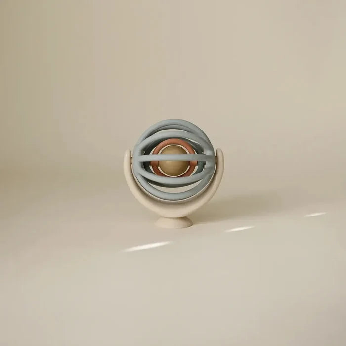A modern spherical baby teether or sensory toy with concentric rings in muted blue-gray, beige, and rose gold, displayed on a matching beige stand against a warm beige background.