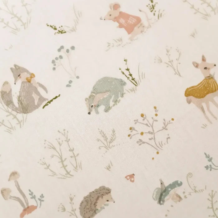 A close-up of a fabric or wallpaper pattern featuring cute woodland animals like a fox, hedgehog, deer, rabbit, and a mouse, with small wildflowers and grass on a warm off-white background.