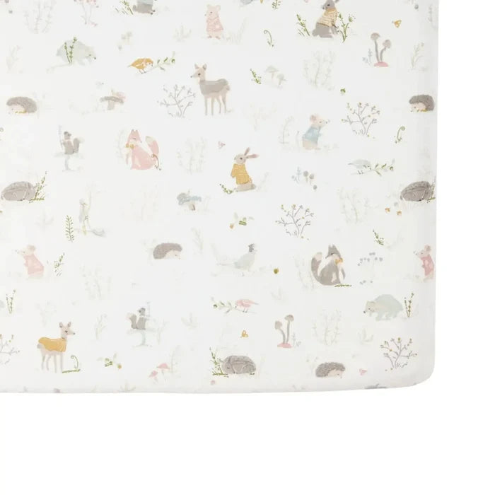 A close-up of a fitted crib bumper or mattress cover featuring a whimsical woodland animal print on a warm off-white background. The pattern includes small illustrations of a deer, rabbit, hedgehog, squirrel, birds, and mushrooms in muted pastel tones of beige, brown, pink, and yellow.