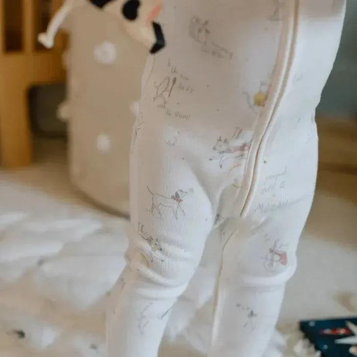 A baby wearing a white footed sleeper with a cute cartoon dog print and a front zipper, standing on a white quilted blanket.
