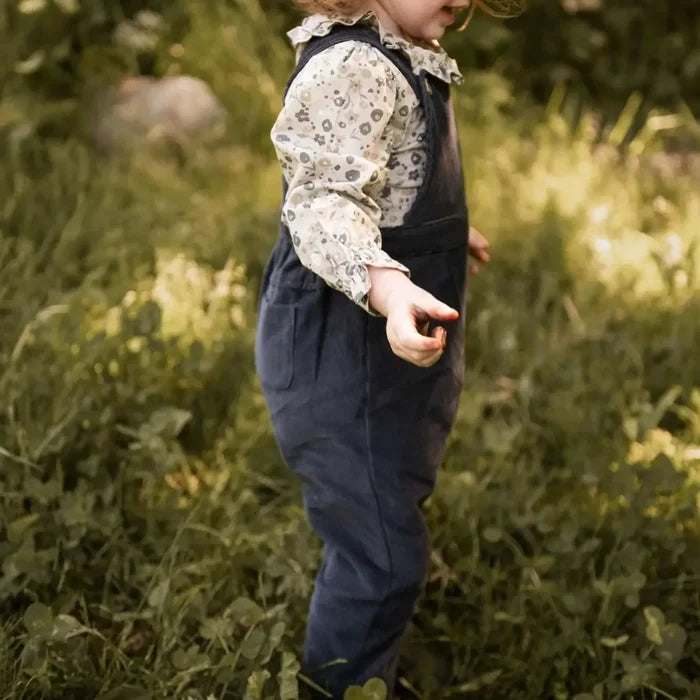 A toddler standing outdoors in tall grass, wearing navy blue overall-style pants over a cream patterned long-sleeve shirt.