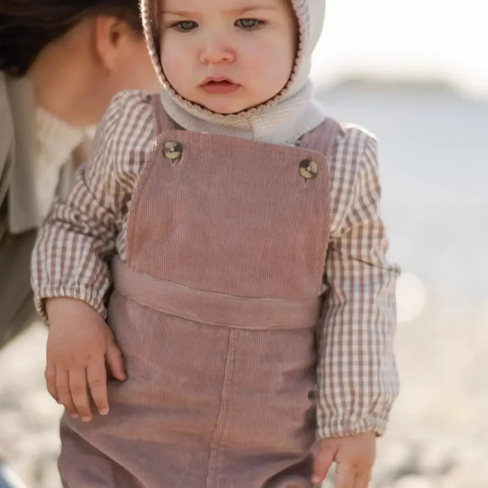 A toddler standing outdoors wearing dusty mauve corduroy overalls over a beige and white gingham long-sleeve shirt, with a cream knit bonnet. A woman is partially visible on the left in a soft-focus outdoor setting.