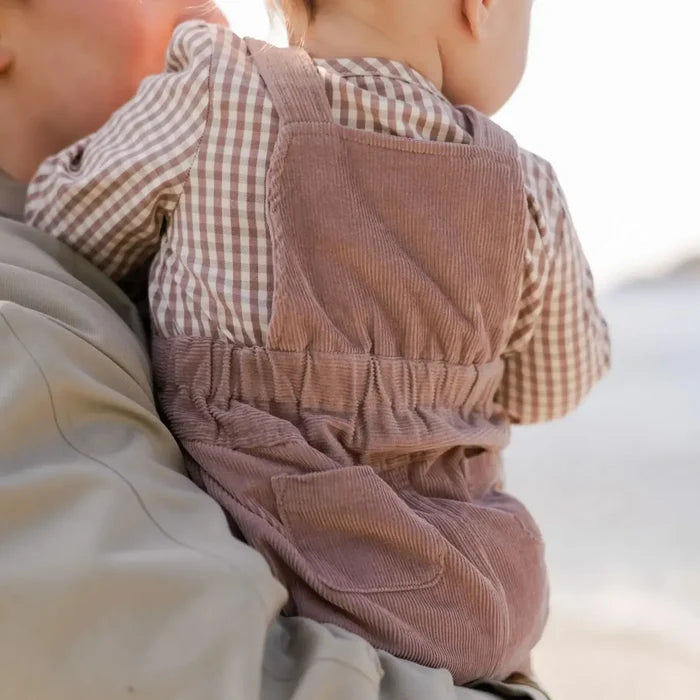A close-up lifestyle shot of a baby wearing dusty rose corduroy overalls and a brown-and-white gingham shirt, being held by an adult outdoors.