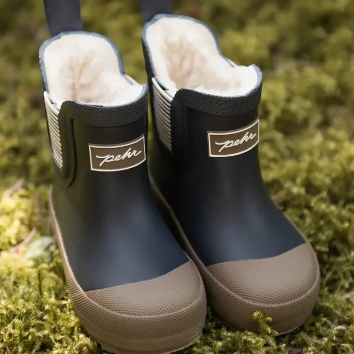 A pair of children's rubber rain boots with a dark navy blue upper, beige toe caps and soles, and a soft cream fleece lining, resting on a bed of green moss.