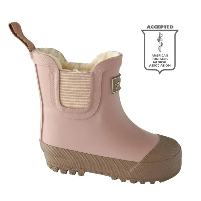 A light pink children's rain boot with a brown sole and pull tab, featuring a striped elastic side panel and a plush cream lining. A shield-shaped 'Accepted' badge from the American Pediatric Medical Association is displayed on the right side of the white background.