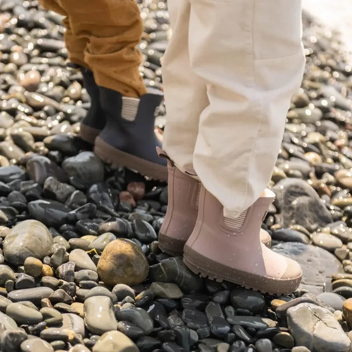 Two pairs of feet wearing pink rubber boots on a pebbly beach.