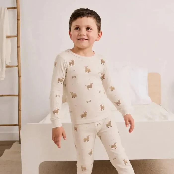 A young boy wearing a cream-colored long-sleeve pajama set with a repeating brown sheep pattern, standing in a brightly lit bedroom.