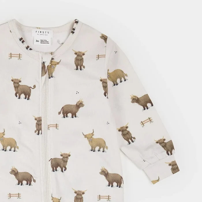 A long-sleeve baby zip-up romper by Firsts London, featuring an all-over print of Highland cows and small wooden fences on a warm off-white background.