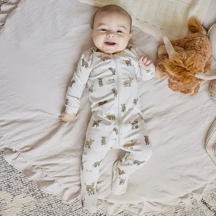 A baby lying on a beige blanket, wearing a footed zip-up sleeper with a brown and gray animal print. A plush toy is visible next to the baby.