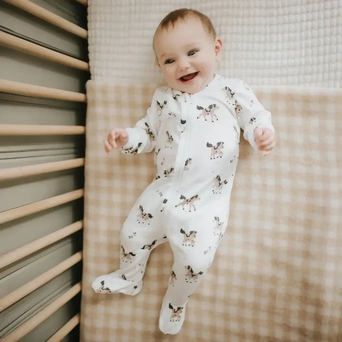 A baby smiling while wearing a white footed sleeper with a brown and black dog print, lying on a beige and white checkered blanket in a crib.