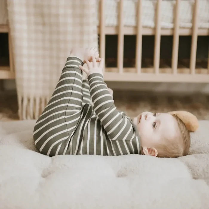 A baby lying on a fluffy cream rug in a nursery, wearing a ribbed long-sleeve one-piece in olive green and white stripes.