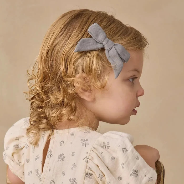 A young child with blonde curly hair wearing a light gray bow headband and a cream-colored floral dress, shown from the back while sitting on a wooden chair against a warm beige background.