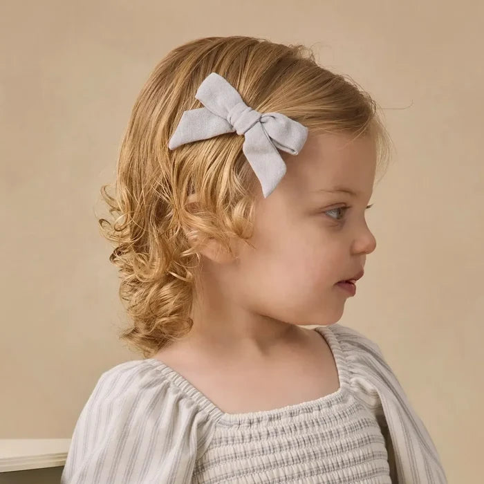 A young girl with blonde curly hair wearing a cream and beige vertically striped smocked dress with puffed sleeves and a matching striped bow headband, leaning on a light-colored wooden bench against a warm beige background.