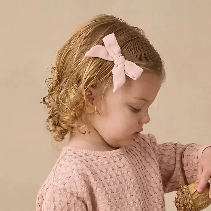 A toddler girl stands in a studio setting wearing a matching dusty pink knit long-sleeve top and shorts set. She has a light pink bow in her hair and is looking down at a small wooden bucket filled with pinecones on a textured cream rug against a warm beige background.