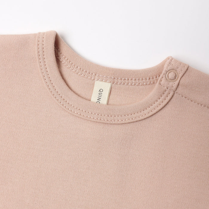 Close-up of a baby or toddler crewneck sweatshirt in a dusty rose pink color, featuring a cream-colored brand tag that reads 'quino' and a snap button on the shoulder.
