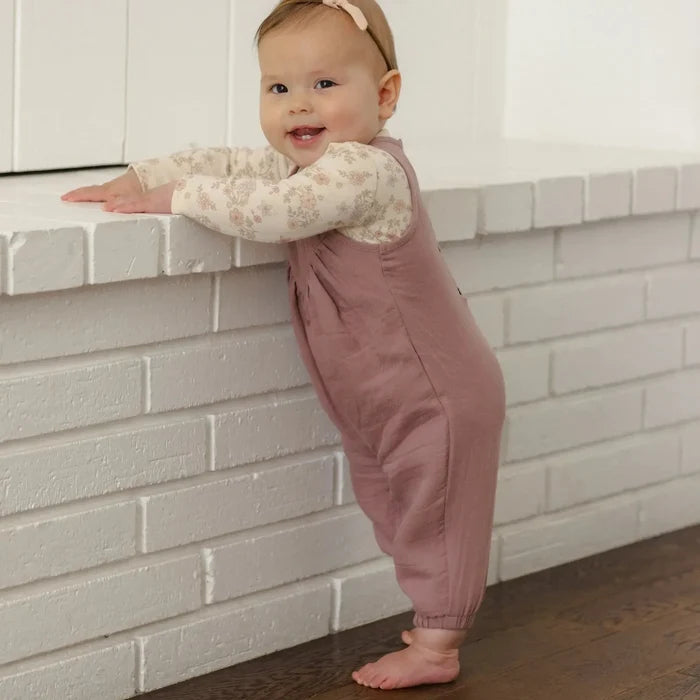 A baby standing on a dark wood floor next to a white brick fireplace, wearing a dusty rose overall jumpsuit over a cream floral long-sleeve shirt and a matching beige headband.
