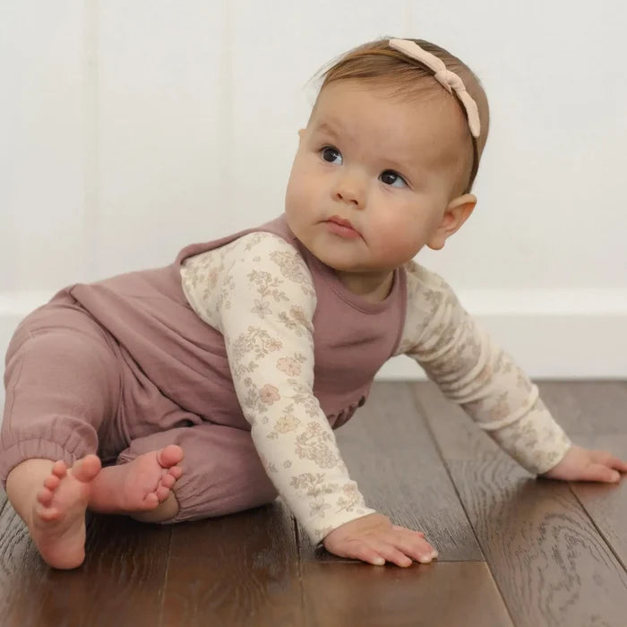 A baby crawling on a dark wood floor against a white paneled wall, wearing a mauve-brown sleeveless jumpsuit over a cream floral long-sleeve shirt and a matching pink headband.
