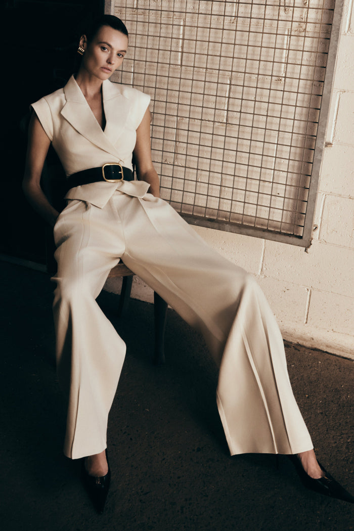 A model wearing natural beige mid-rise wide leg linen pants with a belt, sitting against a tiled wall.