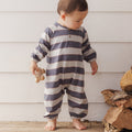 A baby standing barefoot against a white wood-paneled wall, wearing a long-sleeve footless one-piece jumpsuit with horizontal navy and off-white stripes. The baby is holding a small wooden toy.
