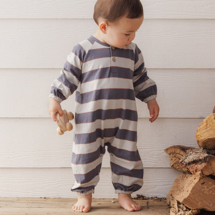 A baby standing barefoot against a white wood-paneled wall, wearing a long-sleeve footless one-piece jumpsuit with horizontal navy and off-white stripes. The baby is holding a small wooden toy.
