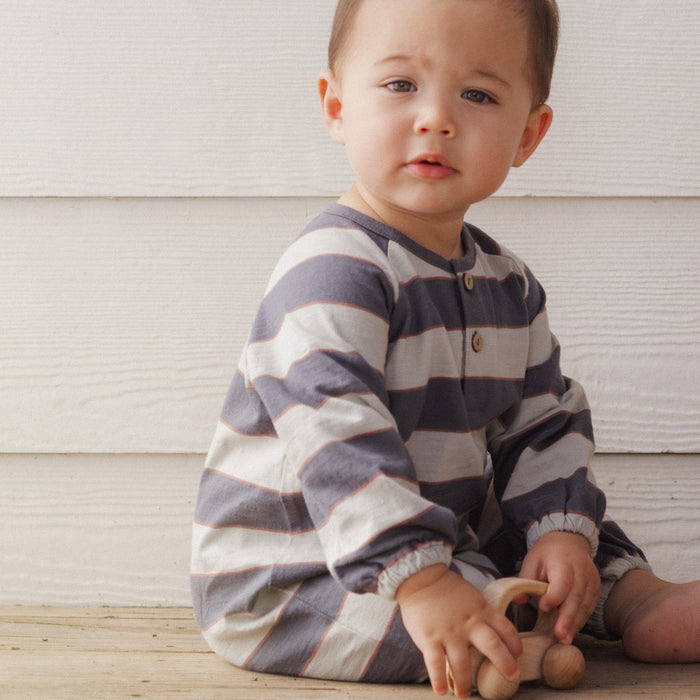 A baby sitting on a wooden floor in front of a white wood-paneled wall, wearing a long-sleeve striped one-piece pajama in navy blue and cream, holding a small wooden toy.