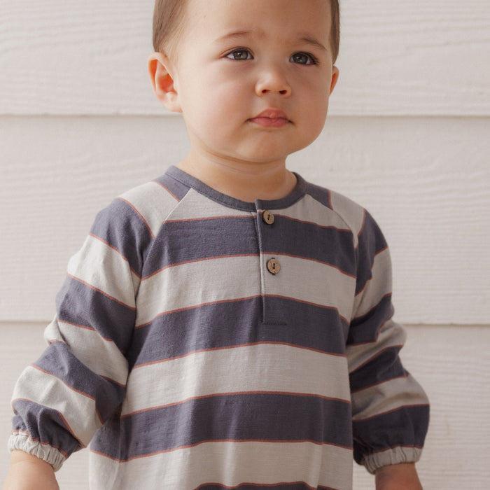 A toddler boy wearing a long-sleeve henley shirt with horizontal navy and beige stripes, standing against a white wood-paneled wall.