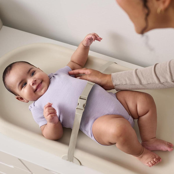 Baby lying on a changing table with a support belt, being held by an adult.