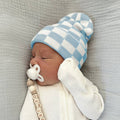Newborn baby wearing a blue and white checkered hat and white outfit with a pacifier on a gray textured surface.