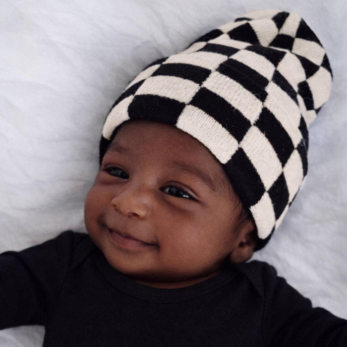A baby lying on a white textured blanket, wearing a black and cream checkered knit beanie and a black long-sleeve bodysuit.