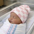 Newborn baby swaddled in a blanket with a pink and white checkered hat in an incubator.