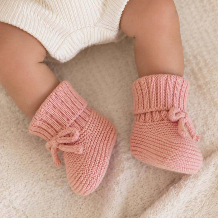 A close-up of a baby's legs wearing soft pink ribbed knit booties with small bows, resting on a textured cream blanket.
