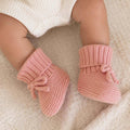 A close-up of a baby's legs wearing soft pink ribbed knit booties with small bows, resting on a textured cream blanket.