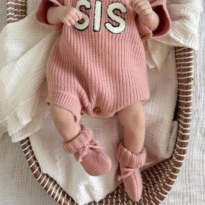 A baby dressed in a pink ribbed romper with 'SIS' lettering, a white bow headband, and pink booties, posed inside a woven oval basket with white muslin fabric.