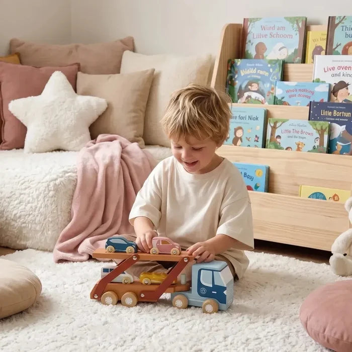 Child playing with a wooden toy car carrier in a cozy living room with books and pillows.
