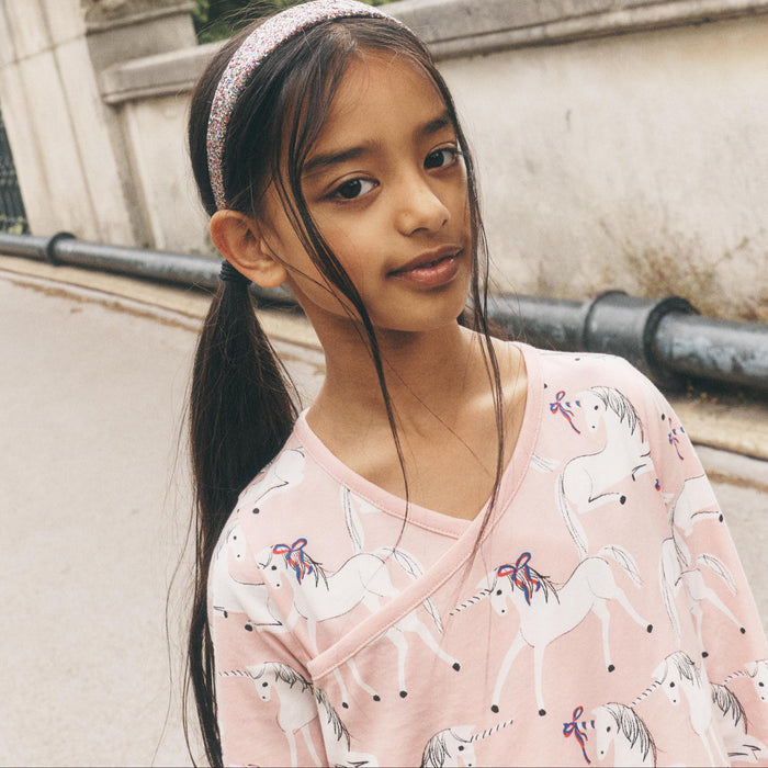 A young girl with dark hair in a ponytail wearing a pink unicorn-print top and a sparkly headband, standing outdoors on a street with a stone wall and buildings in the background.