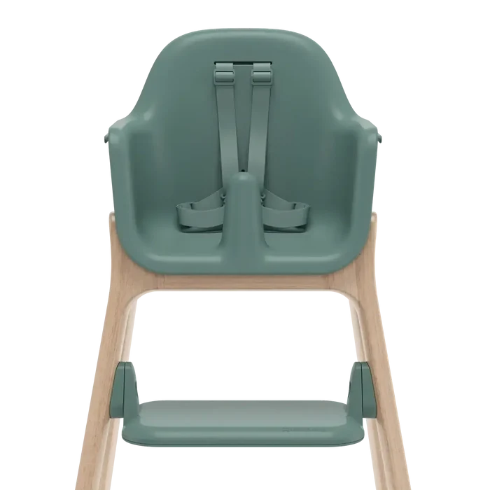 A high chair with a muted sage green molded plastic seat, straps, and footrest, supported by a light natural wood frame, shown from the rear on a white background.
