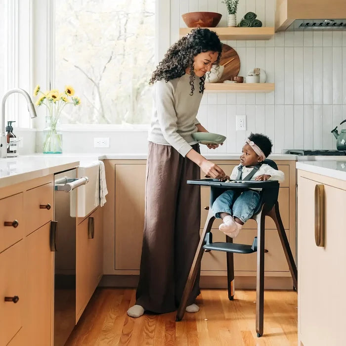 A woman in a beige sweater and brown skirt serves a baby seated in a black high chair in a bright, modern kitchen with light wood cabinets and white subway tile.