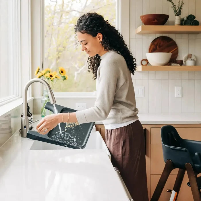 A woman with curly dark hair washes a black sink in a bright, modern kitchen with white countertops and light wood cabinets.