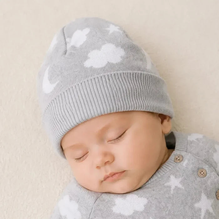 A sleeping newborn baby wearing a light gray knit beanie and matching long-sleeve bodysuit, both decorated with a white cloud and star pattern, lying on a soft cream-colored blanket.