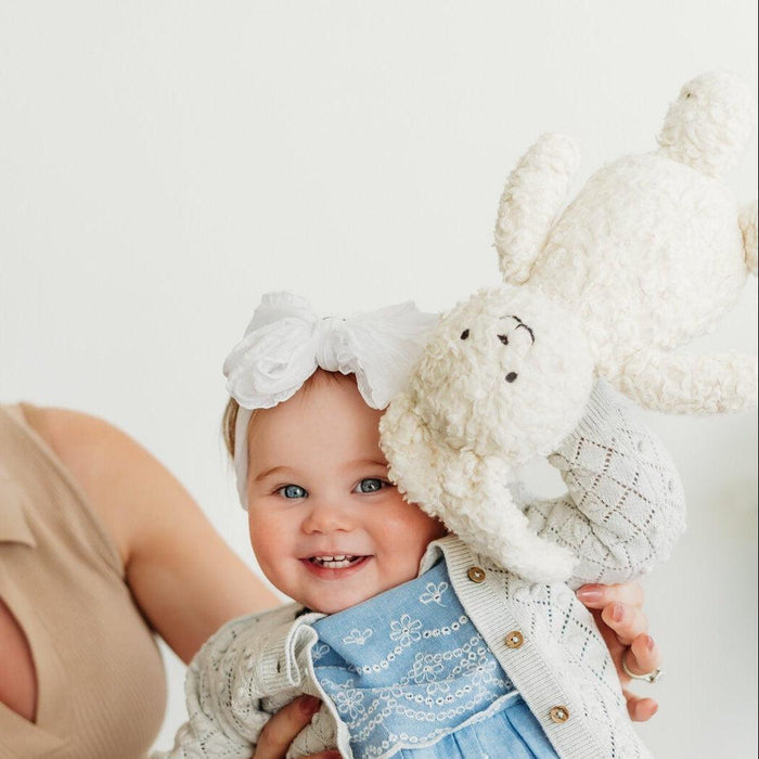 A baby girl wearing a light blue embroidered dress and a white hooded bunny hat is being held by a smiling woman. The baby is also holding a large white plush bunny toy.