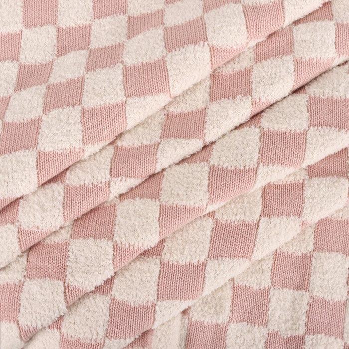 A close-up of a soft woven fabric with a pink and off-white checkerboard pattern, folded diagonally to show the texture and drape.